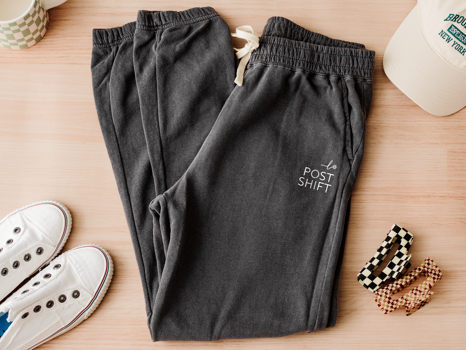 Dark gray sweatpants with 'POST SHIFT' text on a wooden surface with shoes and accessories.