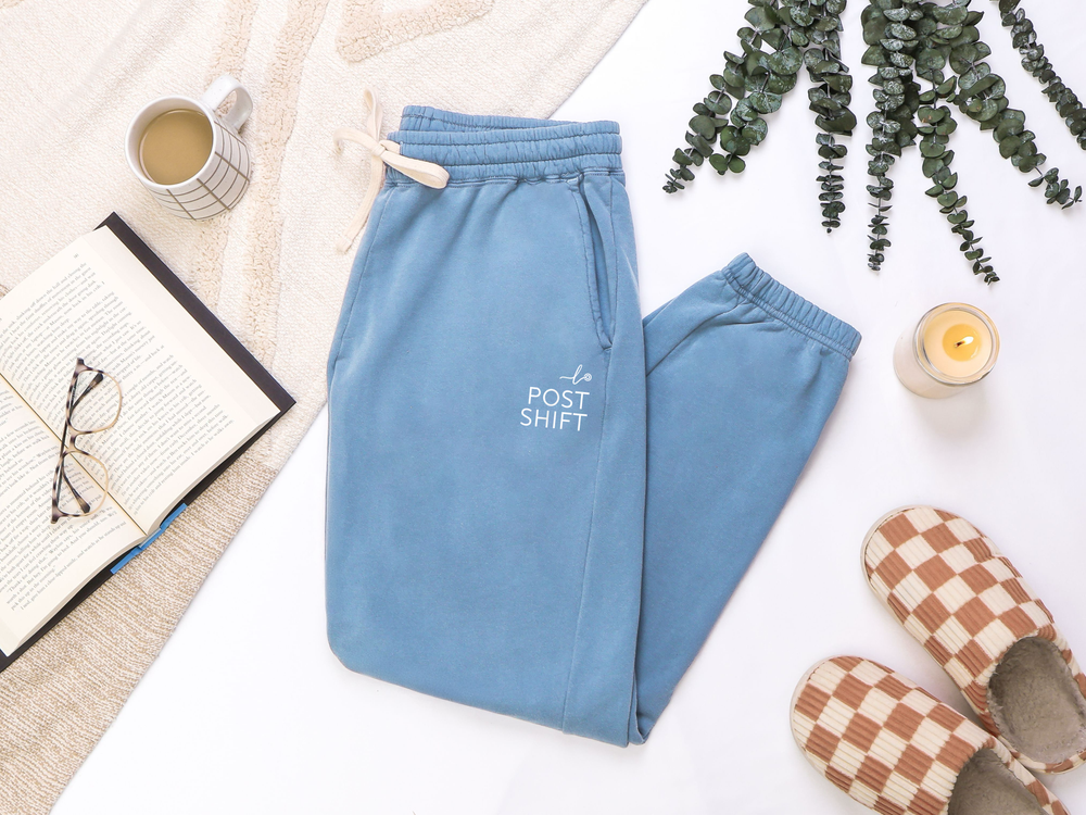 Blue sweatpants with 'Post Shift' text on a white surface with a cup, book, and slippers.