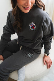 Person wearing a dark gray sweater and pants with a logo, sitting on a white surface.