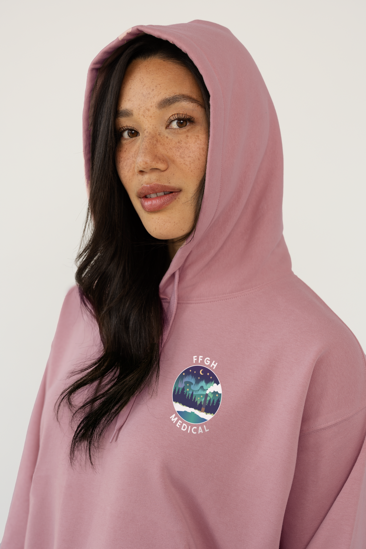 A woman wearing a dusty pink pullover hoodie with a circular chest graphic that reads 'FFGH MEDICAL' around an illustrated night-sky scene, photographed against a clean light gray studio background.